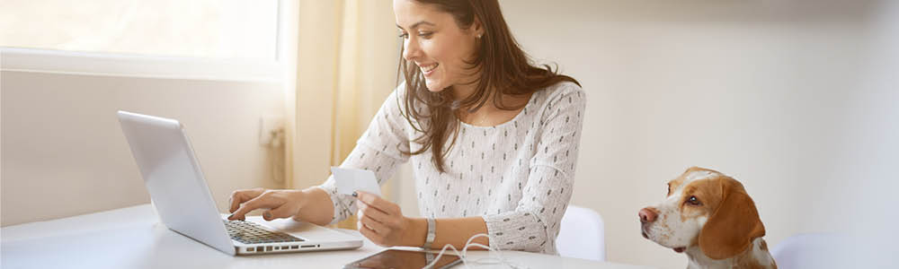 A student using a laptop computer to participate in an online class.  A dog is sitting next to the student looking towards computer screen.